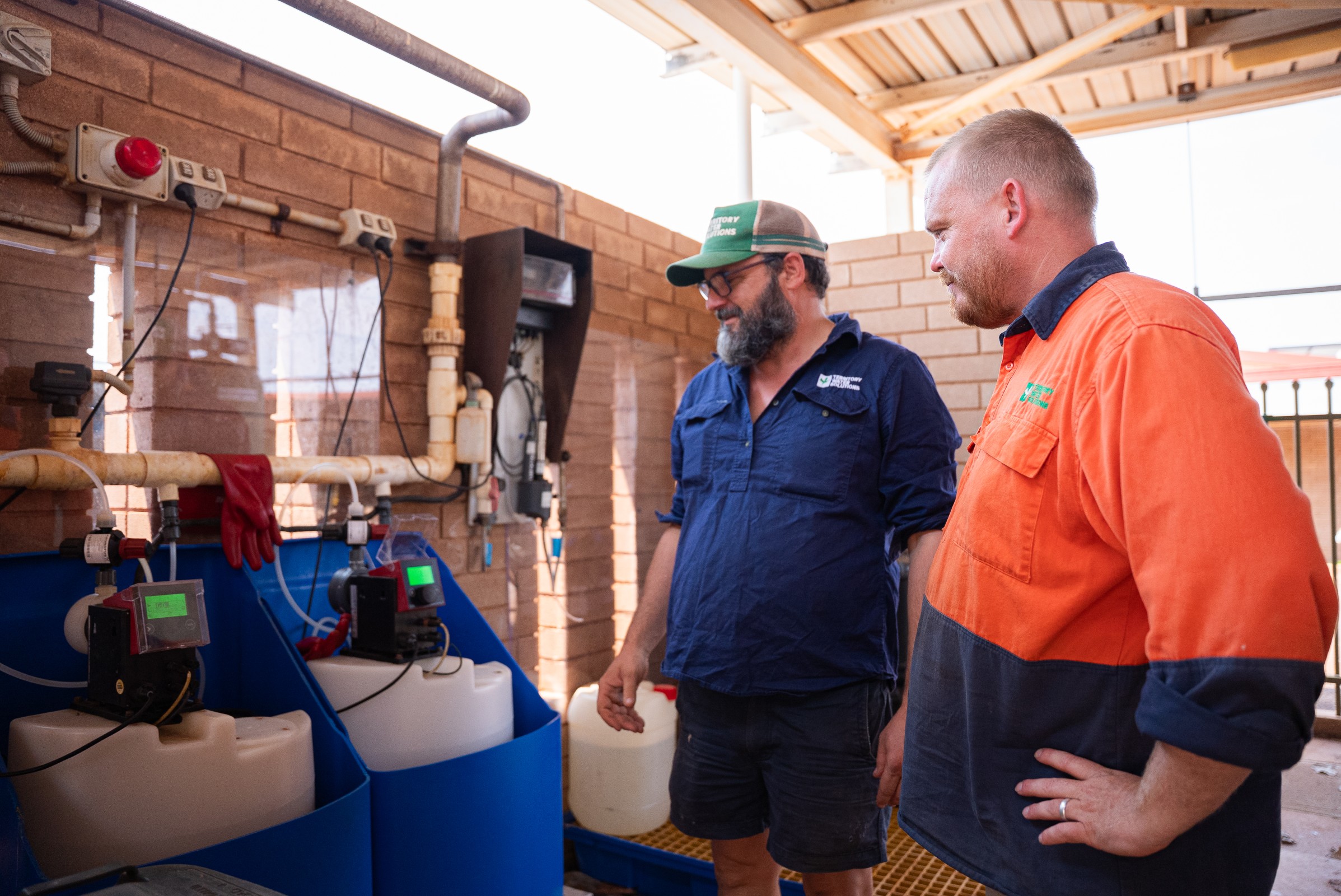 Two men in work uniforms standing inside a facility, inspecting industrial chemical tanks and equipment mounted on a brick wall.