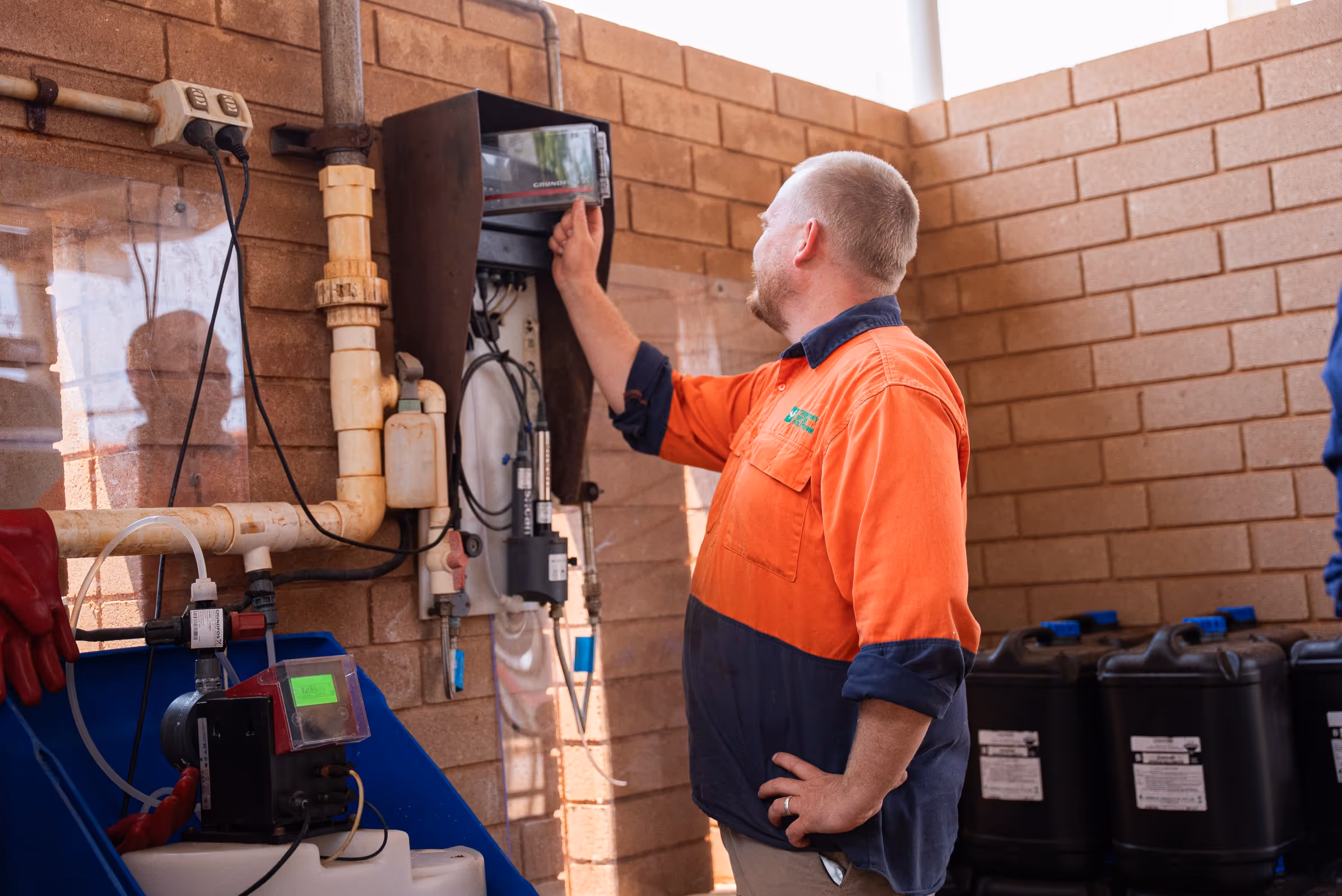Man in orange and navy work shirt adjusting a control panel mounted on a brick wall in an industrial setting.