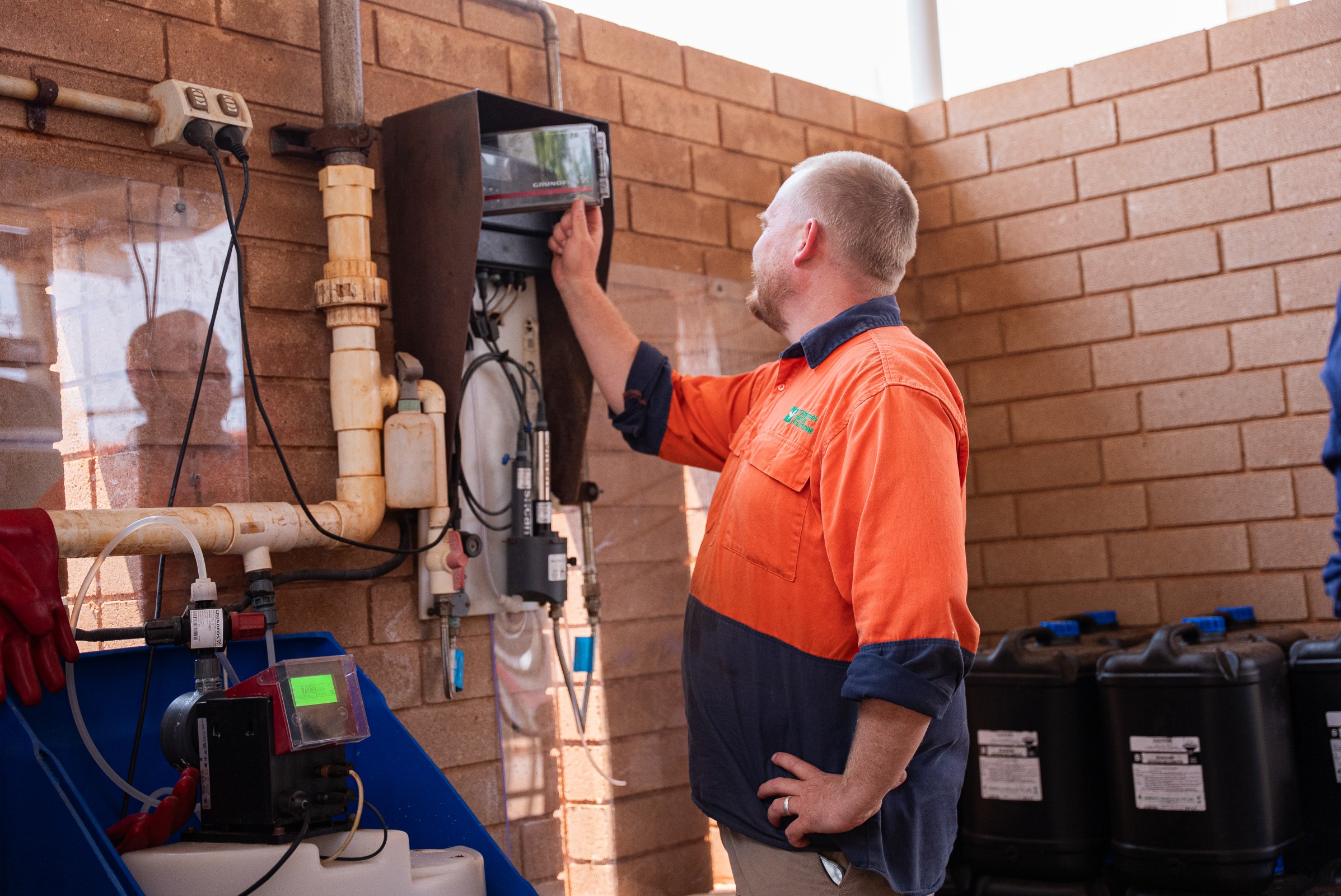 Man in orange and navy work shirt adjusting a control panel mounted on a brick wall in an industrial setting.