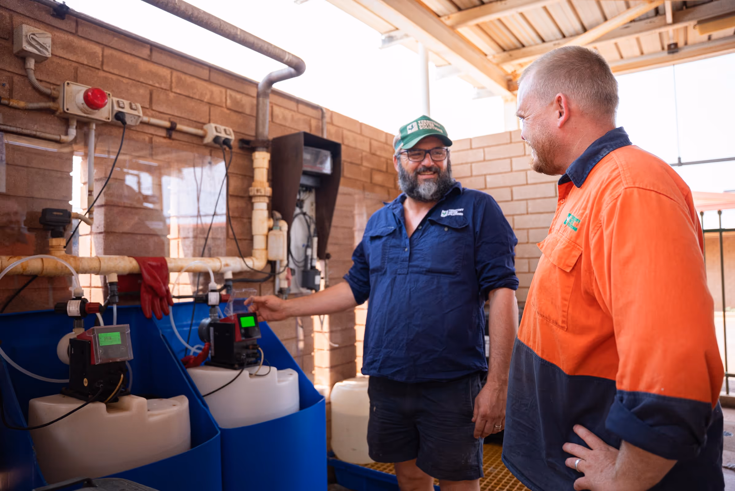 Two men in work uniforms smiling and discussing near industrial equipment with pipes and tanks.