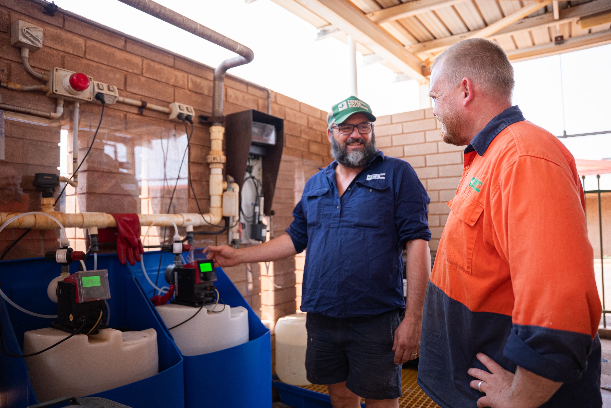 Two men in work uniforms smiling and discussing near industrial equipment with pipes and tanks.