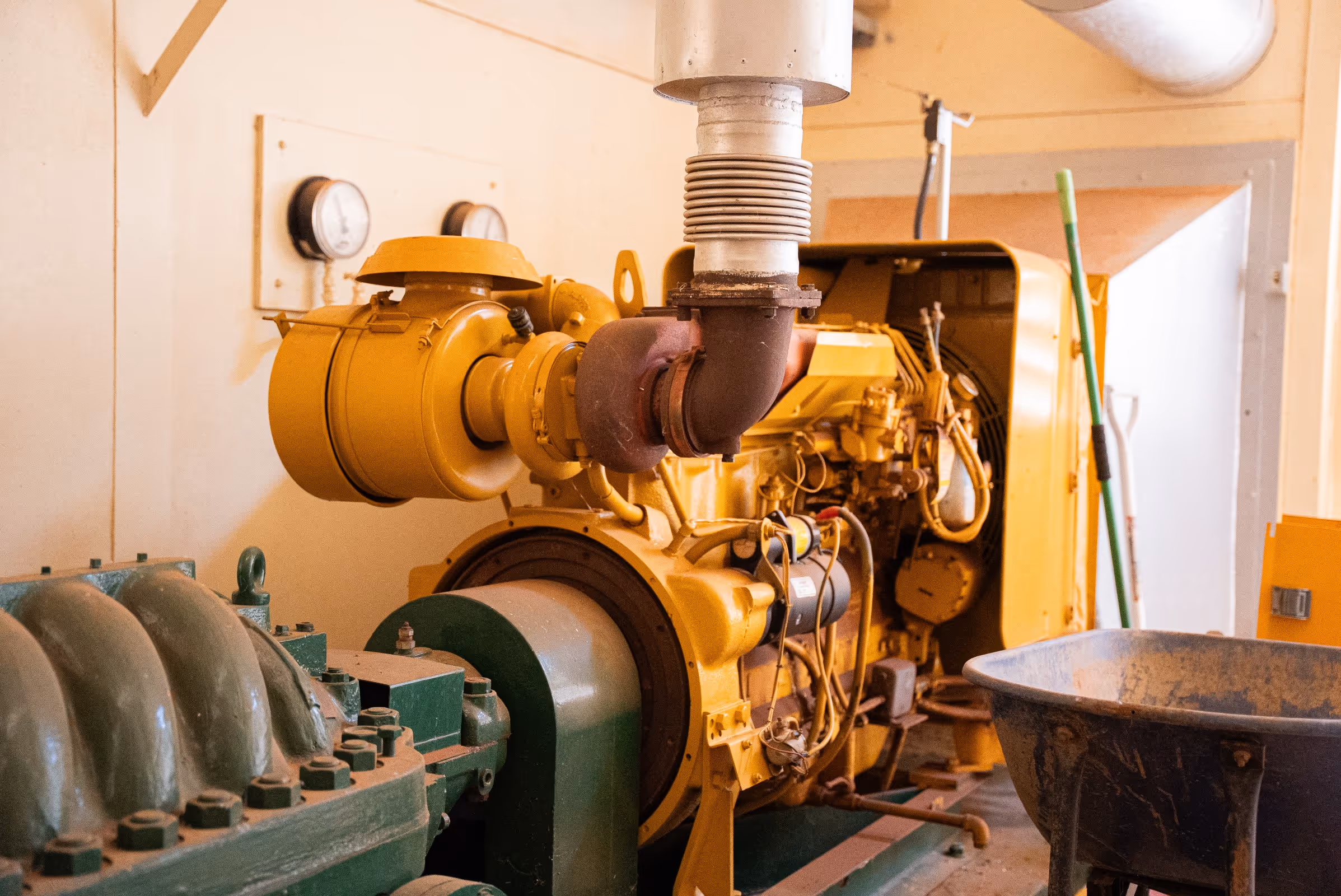 Yellow industrial generator machine with adjacent green equipment inside a utility room.