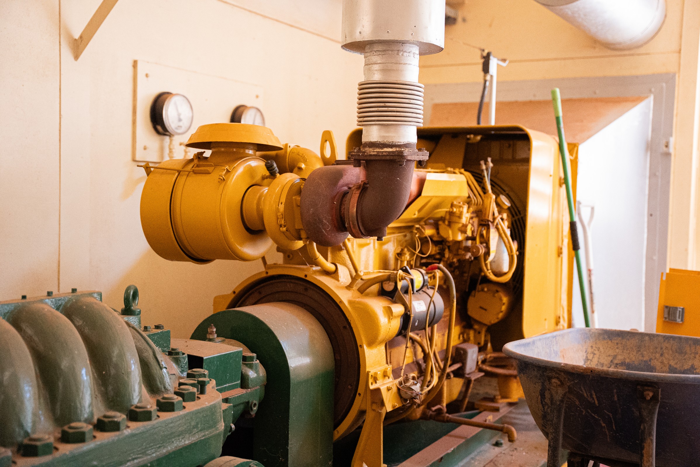 Yellow industrial generator machine with adjacent green equipment inside a utility room.