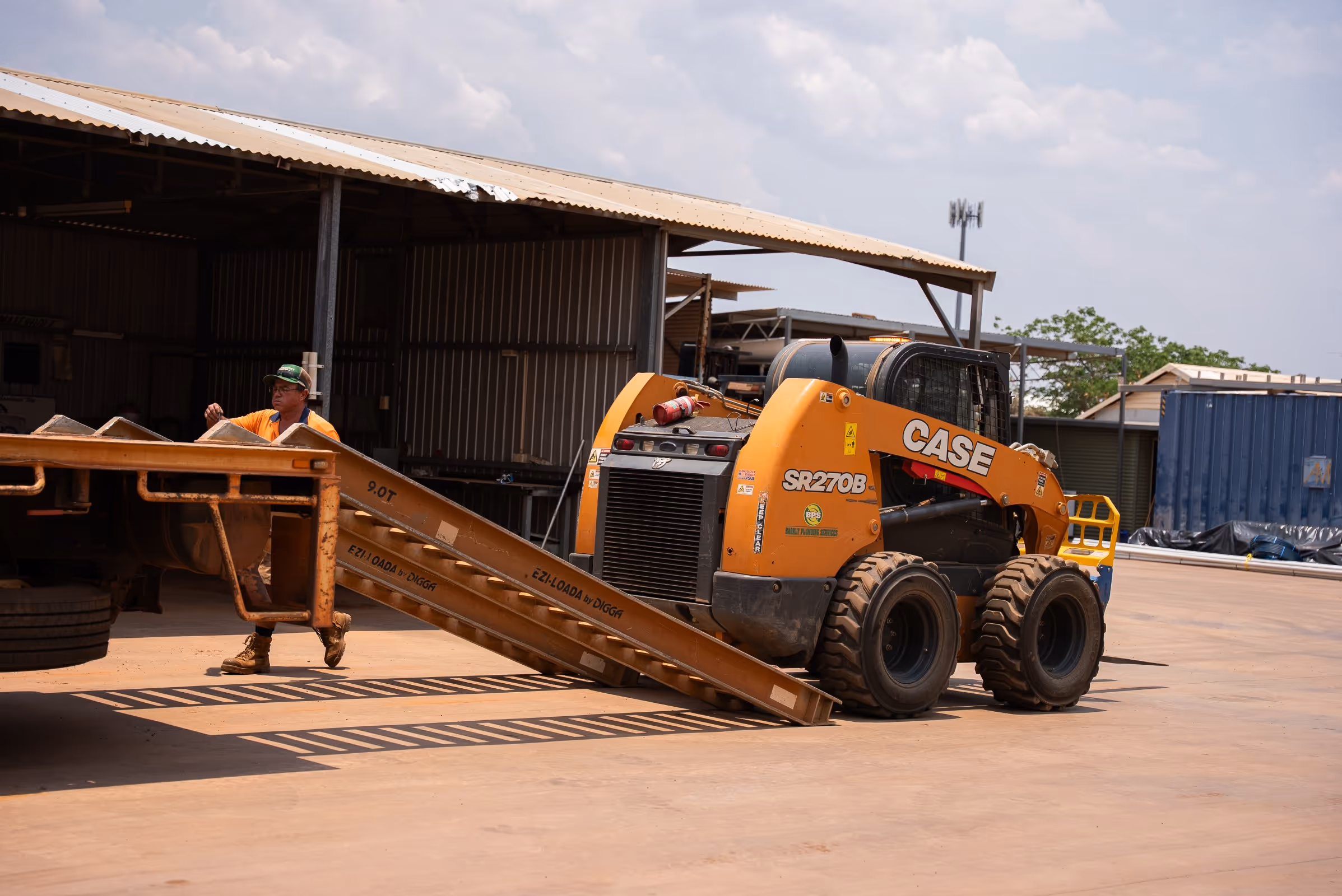 Orange CASE SR270B skid steer loader near a metal ramp with a man in an orange shirt and green hat in the background.