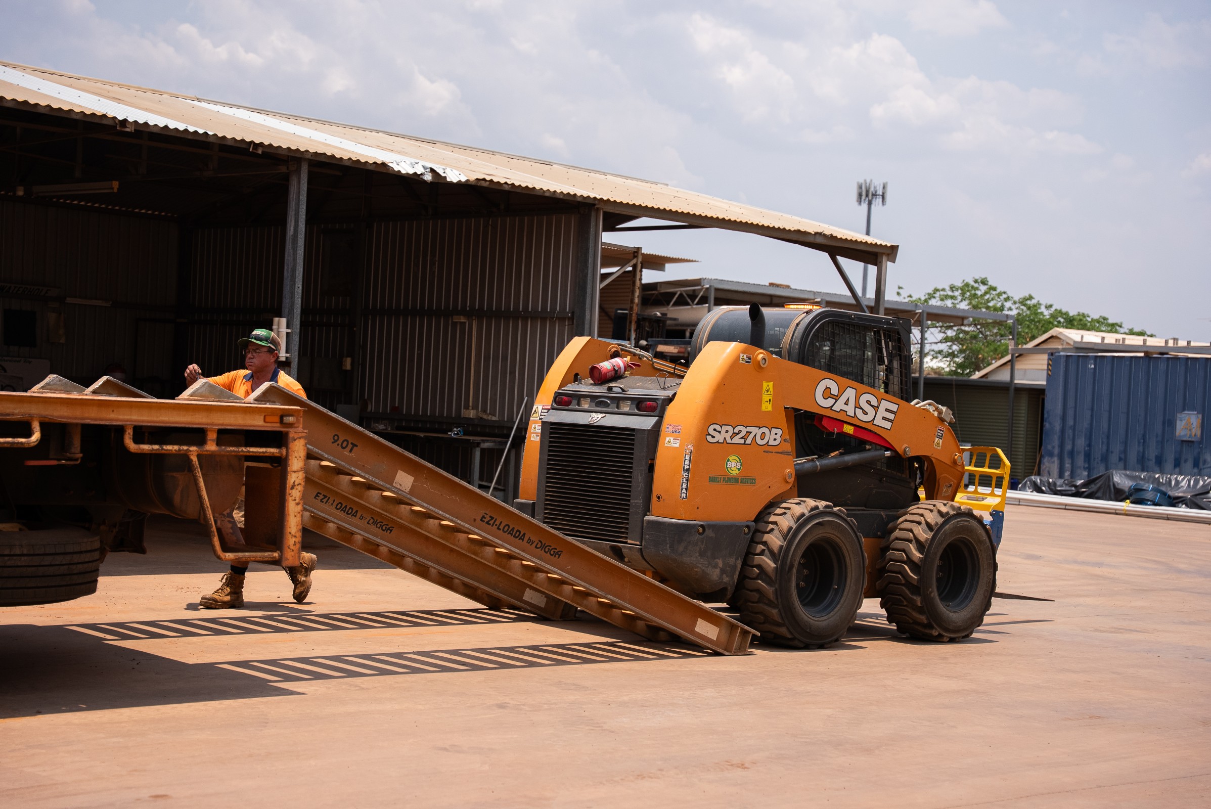 Orange CASE SR270B skid steer loader near a metal ramp with a man in an orange shirt and green hat in the background.