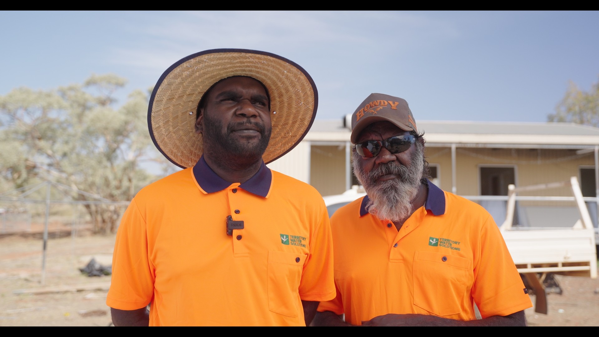Two men wearing bright orange polo shirts with Territory Water Solutions logos, standing outdoors; one wears a wide-brimmed hat, the other a cap and sunglasses.