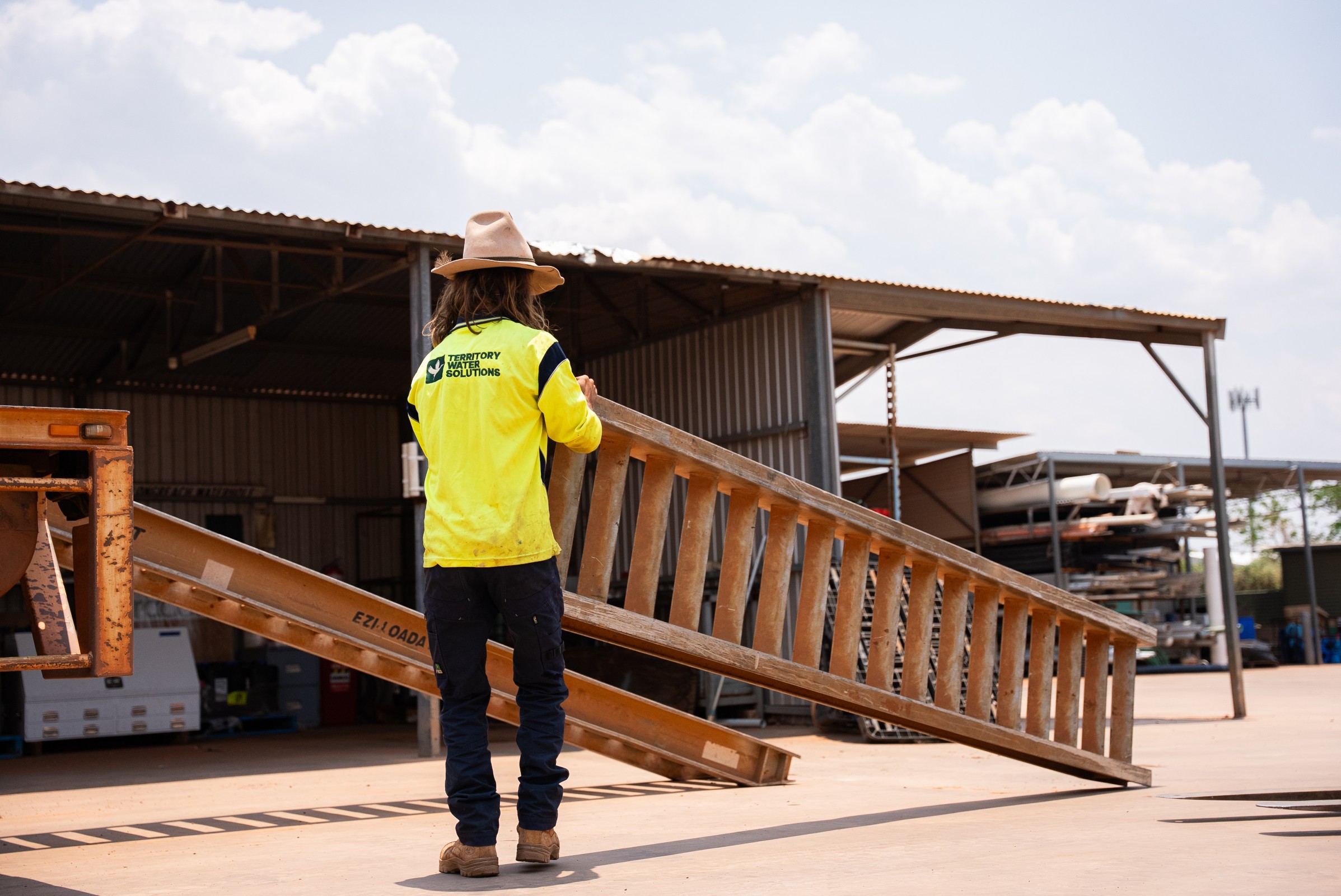 Worker wearing a wide-brimmed hat and high-visibility shirt carrying a wooden ladder at a construction or industrial site.