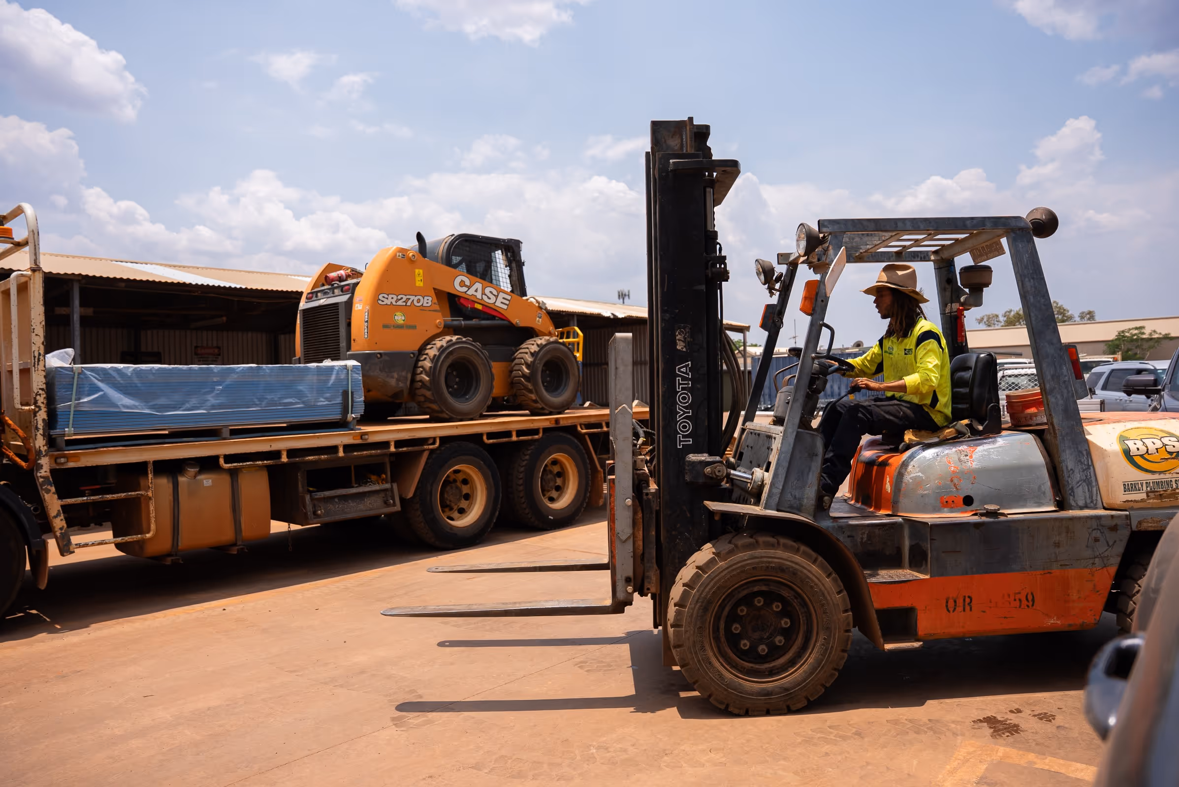 Worker wearing a hat and high-visibility shirt operating a Toyota forklift near a flatbed truck carrying a CASE skid steer loader and wrapped materials.