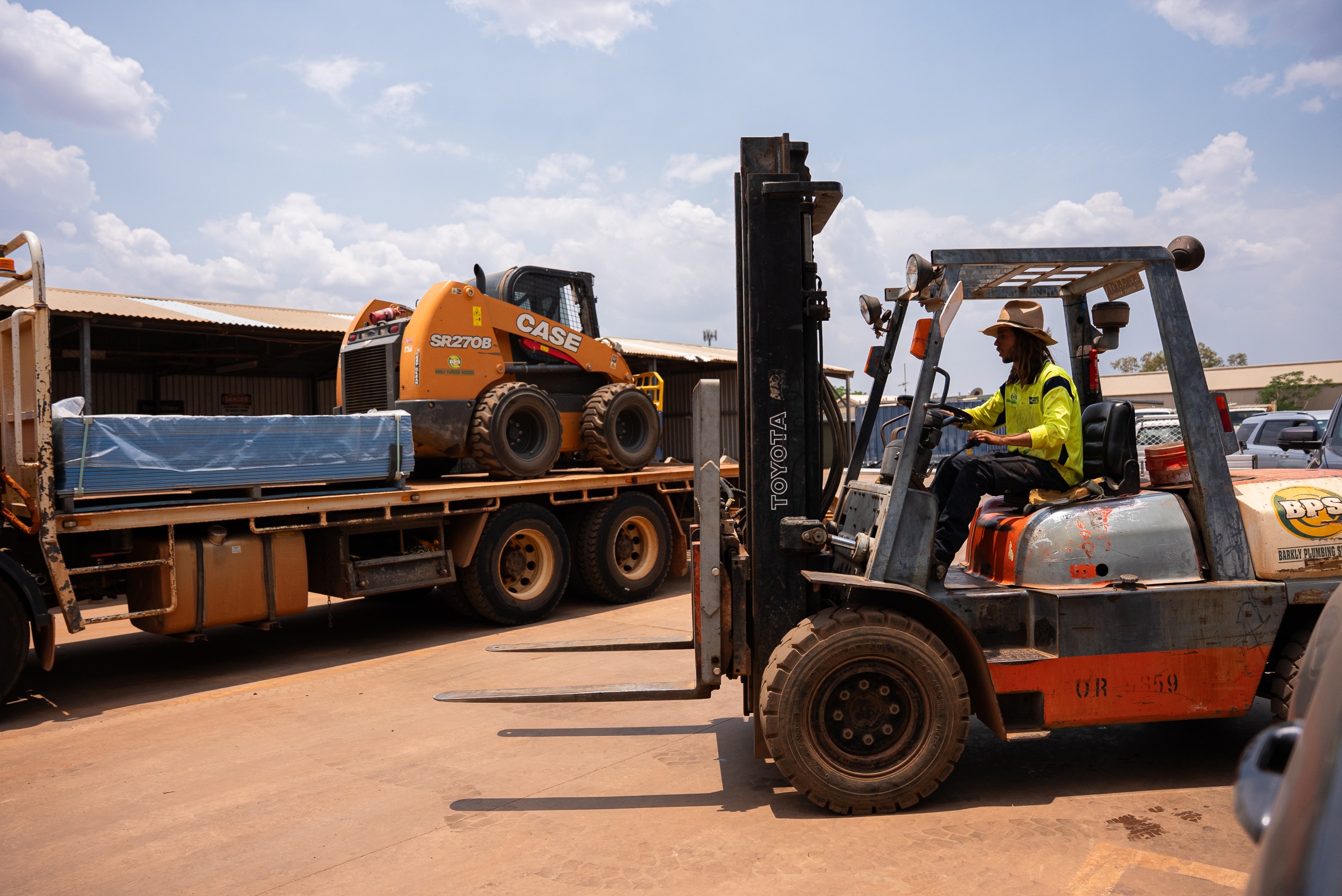 Worker wearing a hat and high-visibility shirt operating a Toyota forklift near a flatbed truck carrying a CASE skid steer loader and wrapped materials.