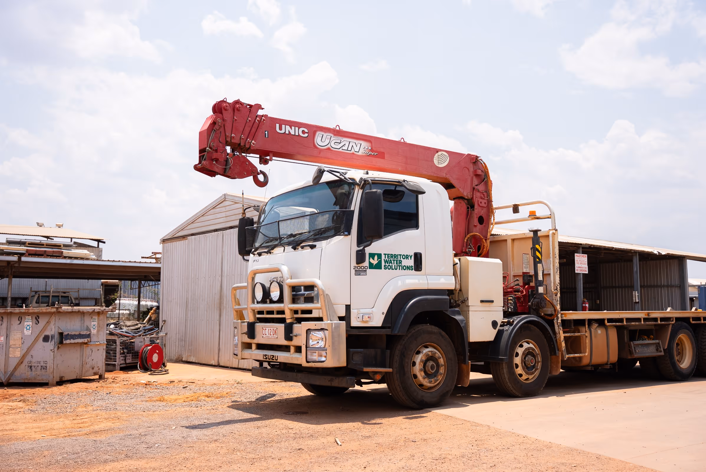 White Isuzu truck with a red UNIC UCAN crane arm parked on a dirt lot beside metal storage sheds.