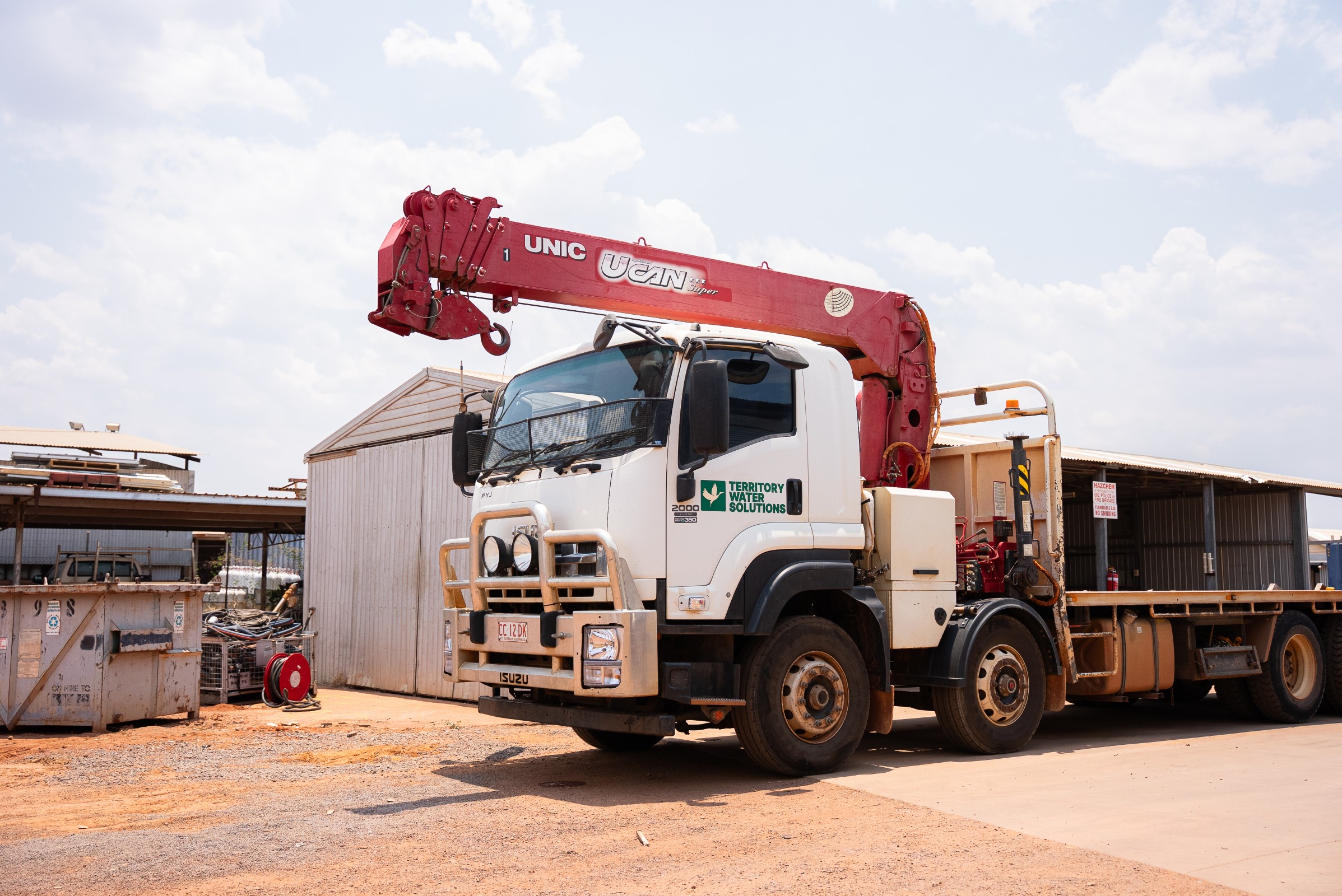 White Isuzu truck with a red UNIC UCAN crane arm parked on a dirt lot beside metal storage sheds.