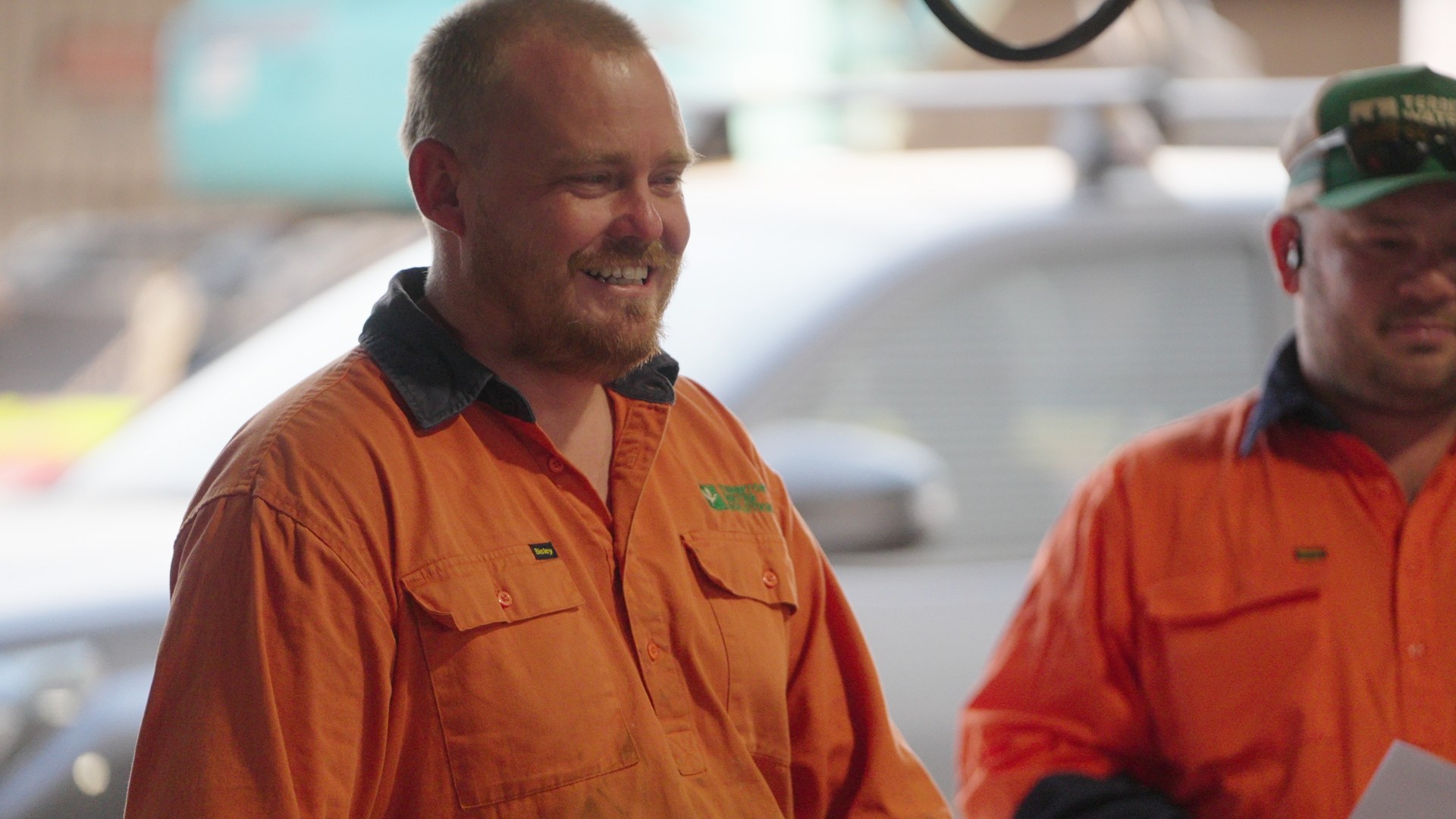 Two men in orange work shirts smiling, one with a beard and the other wearing a green cap.