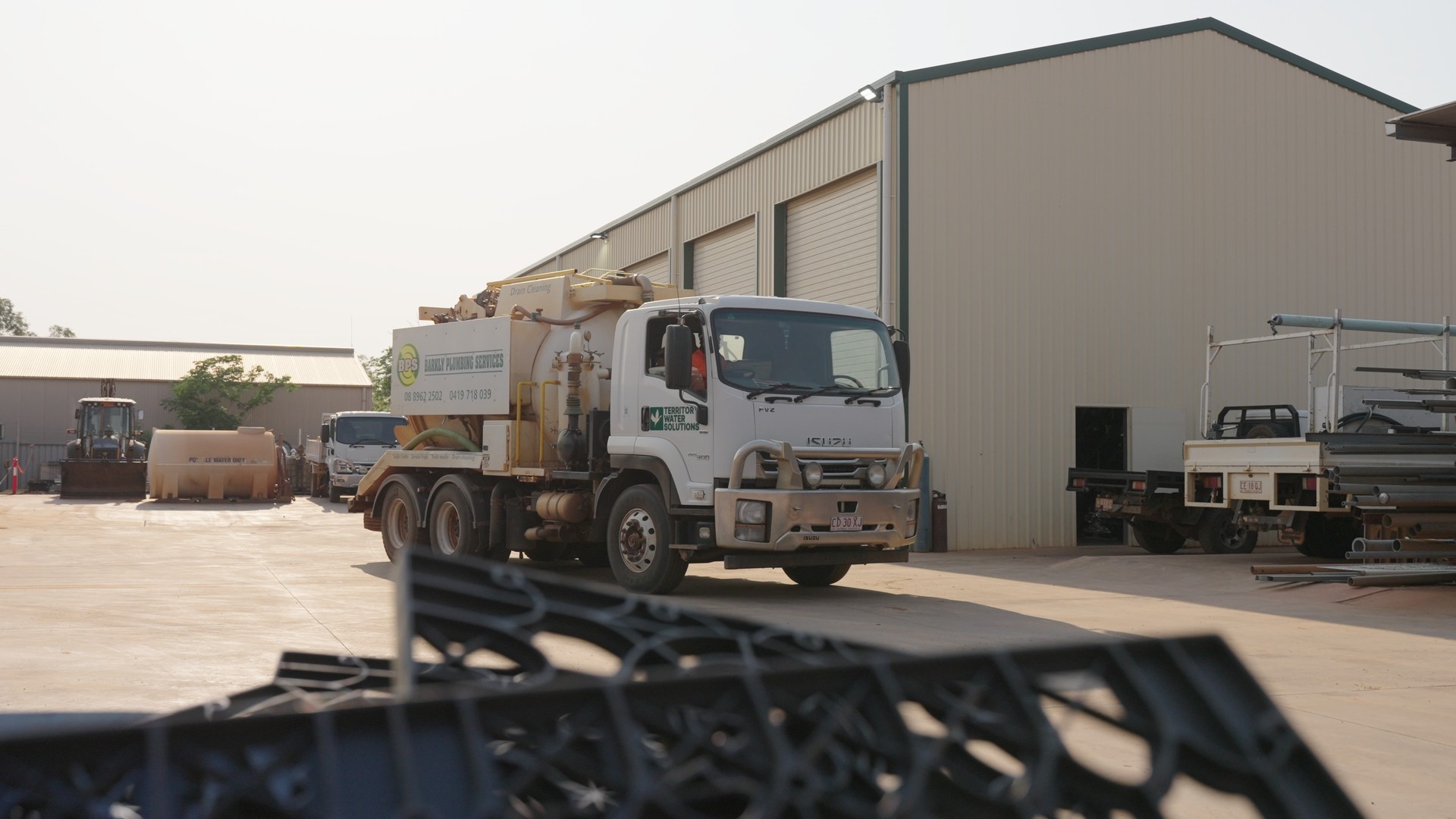A white Isuzu drain cleaning truck parked in an industrial yard with metal buildings and other vehicles in the background.