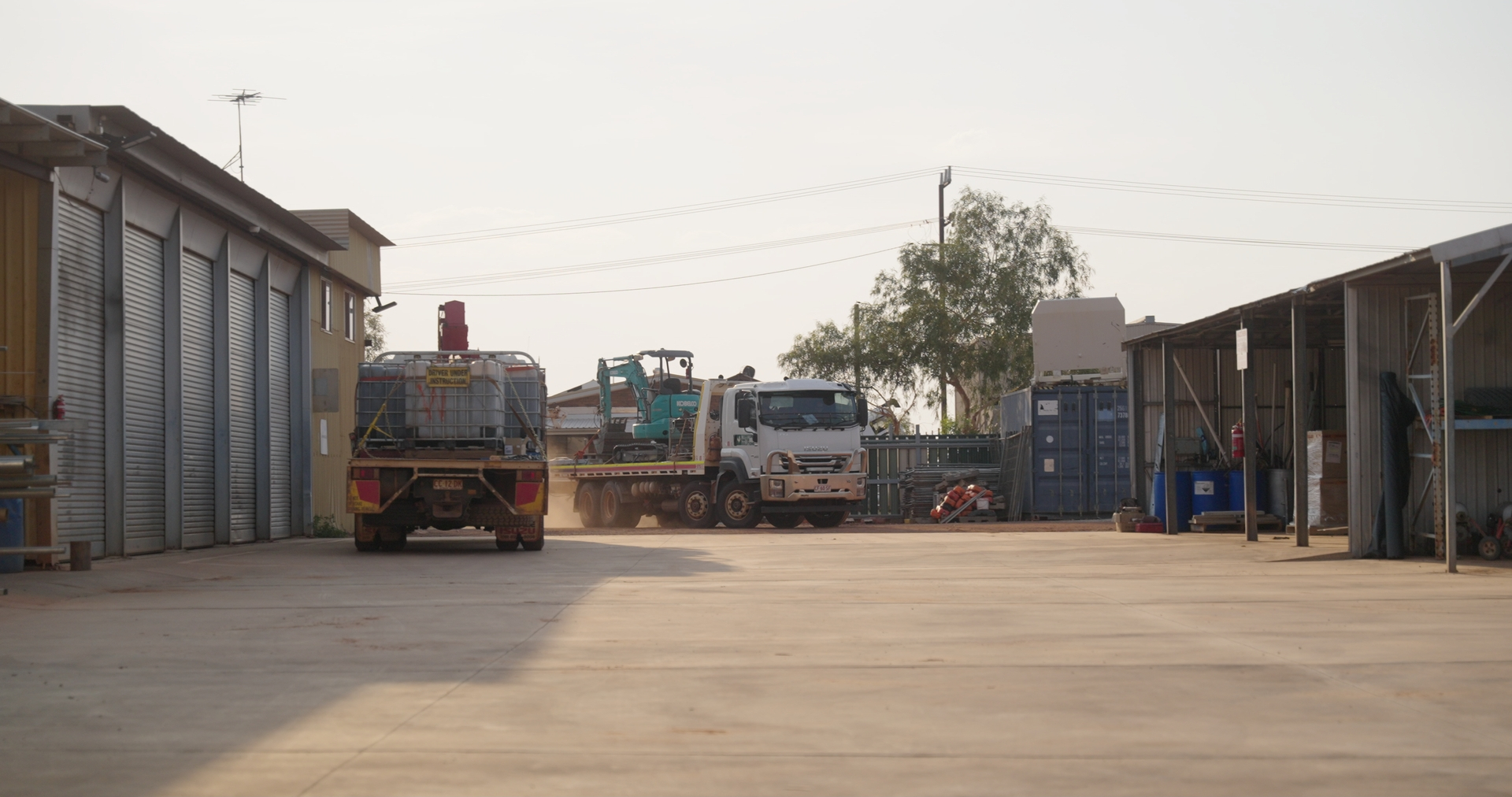 Industrial yard with trucks, one carrying a large water tank and another with a small excavator, surrounded by storage sheds and equipment.