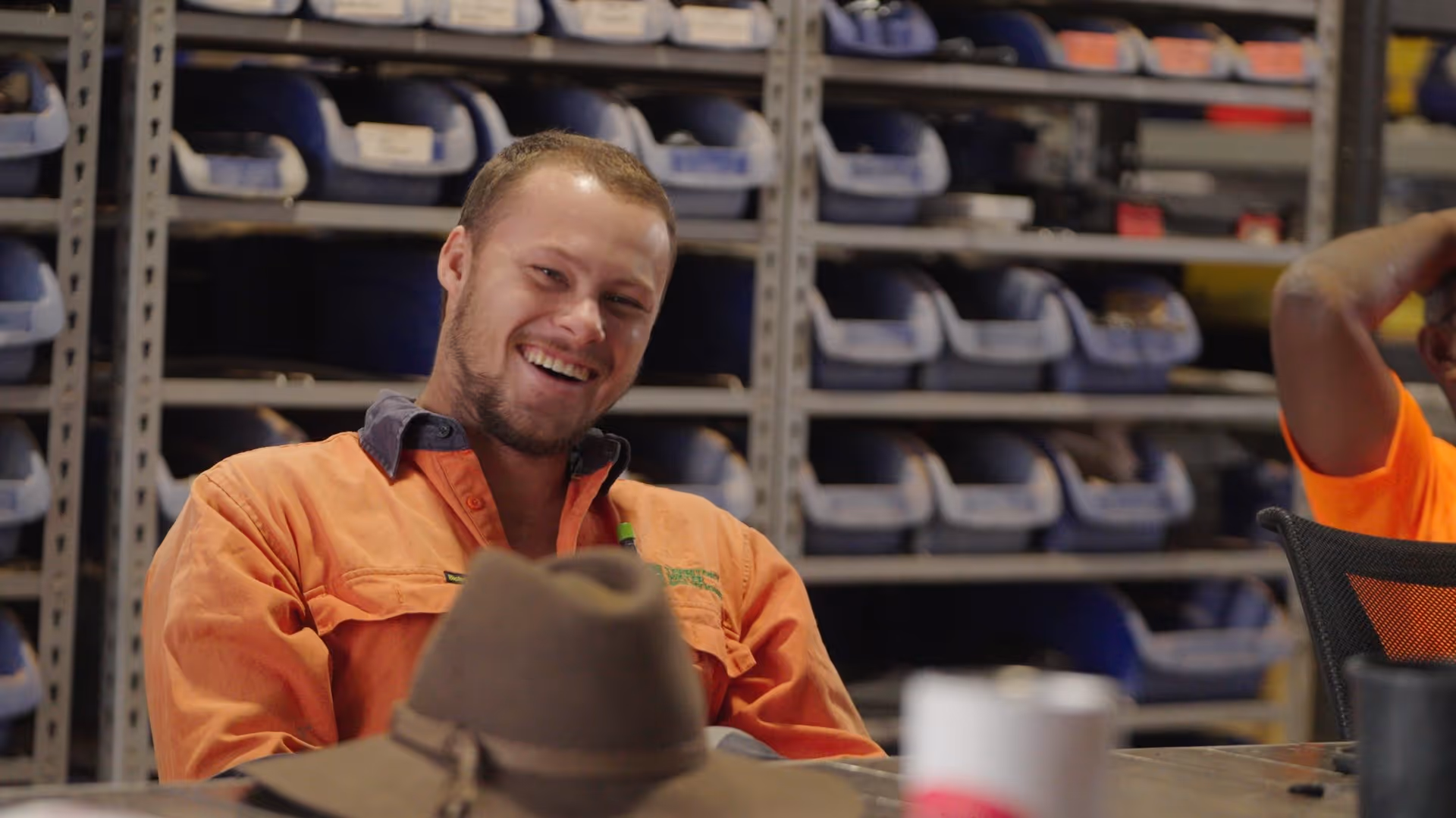 Smiling man in orange work shirt sitting at a table with a brown hat in front and shelves with blue bins in the background.