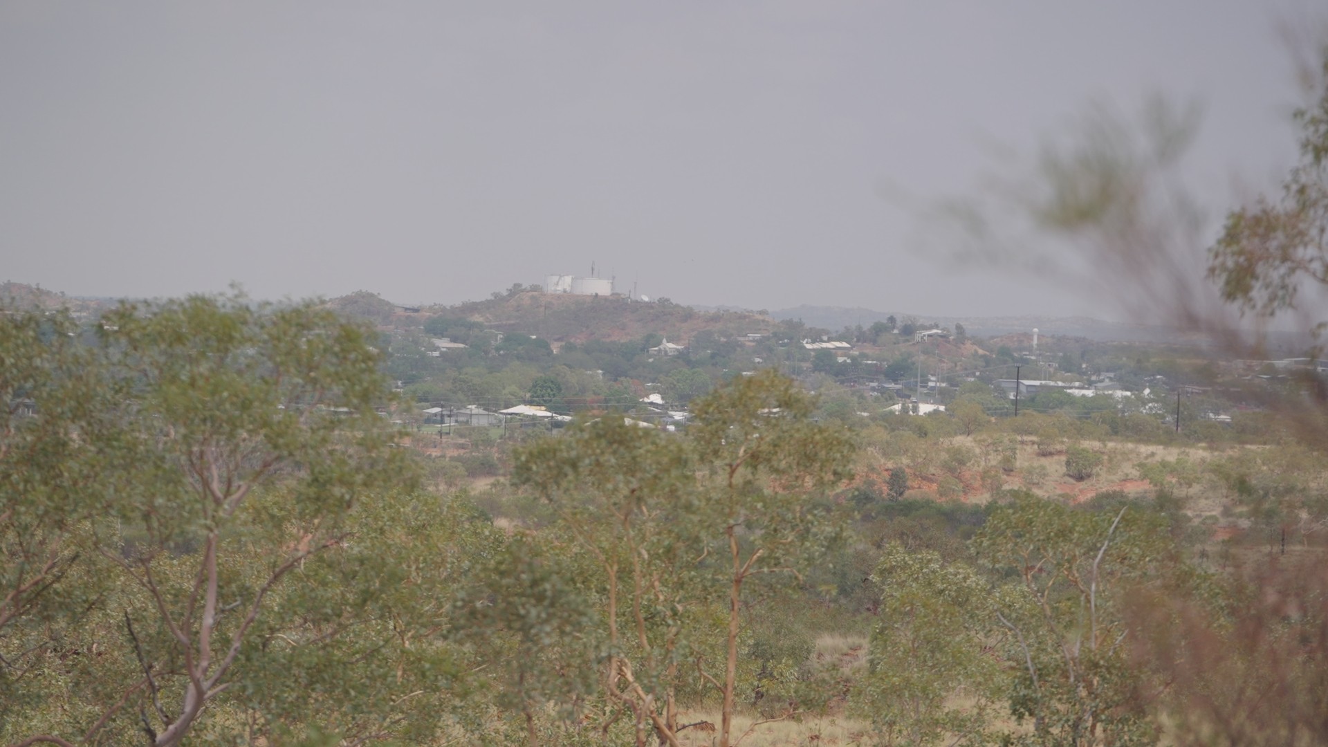 View of a town with scattered buildings and trees, hills in the background under a hazy sky.