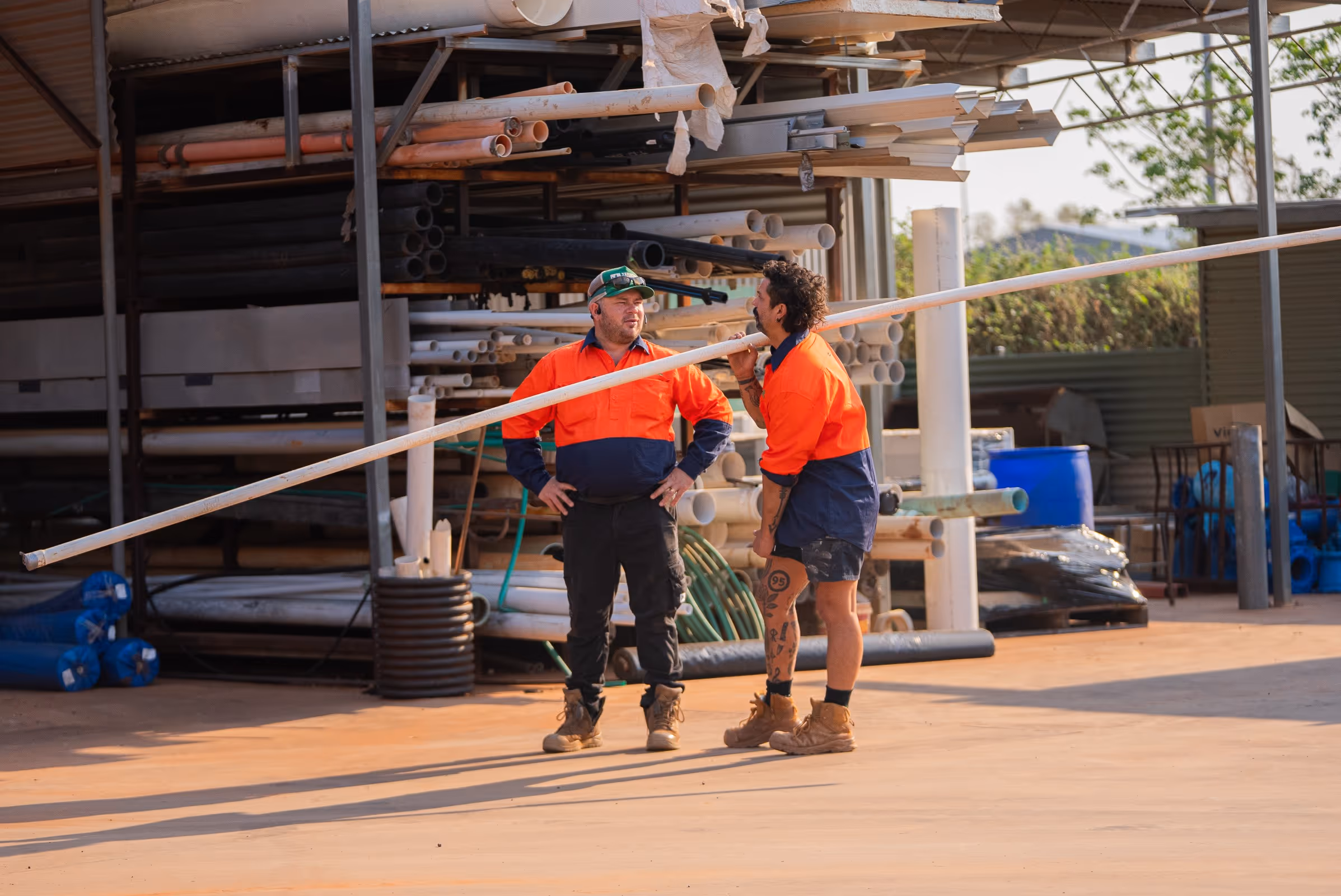 Two construction workers in orange and navy uniforms standing and holding a long white pipe in a storage area with stacked pipes.