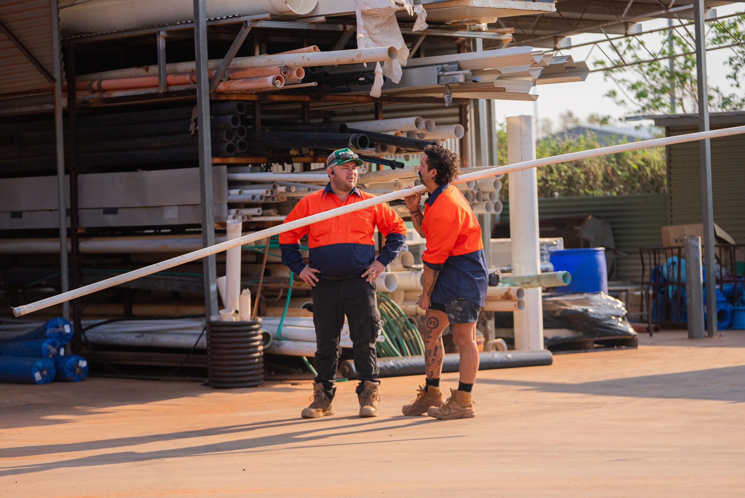 Two construction workers in orange and navy uniforms standing and holding a long white pipe in a storage area with stacked pipes.