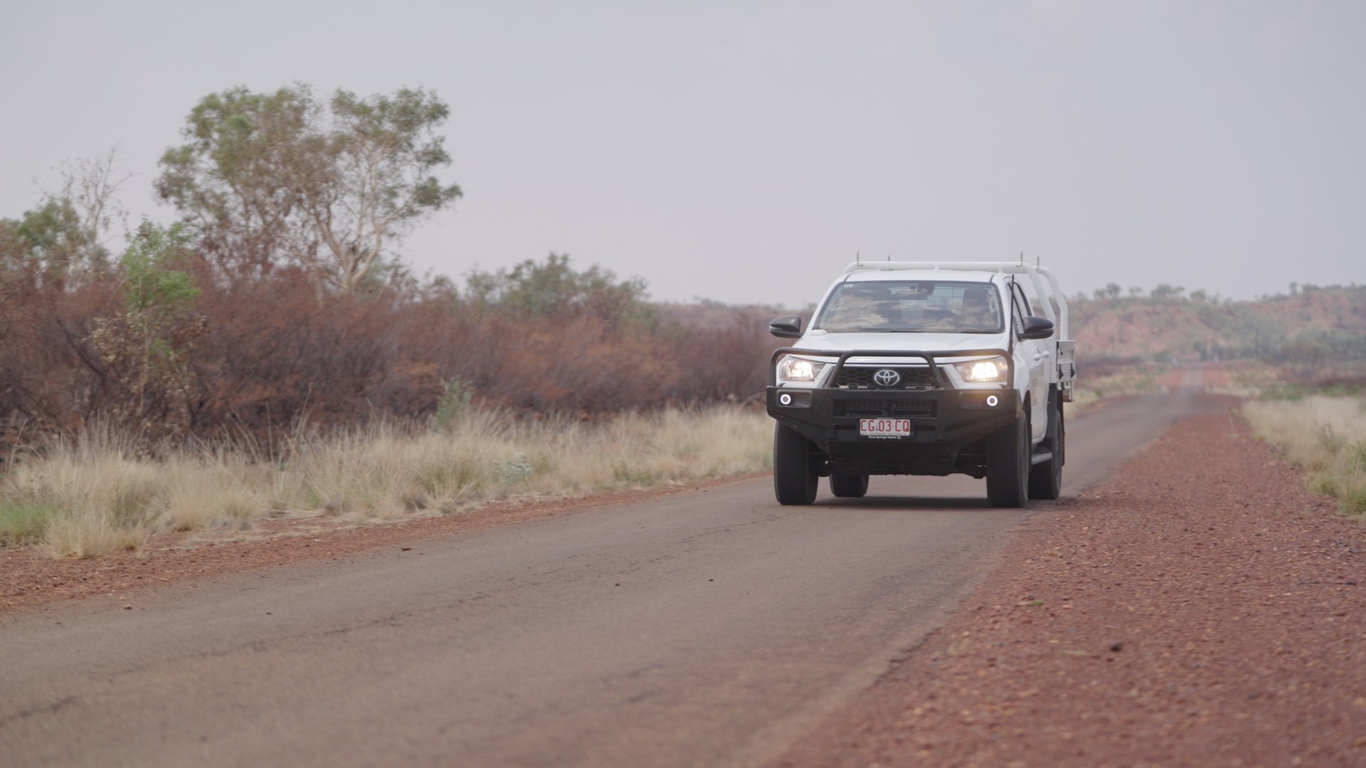 White Toyota pickup truck driving on a rural paved road with dry vegetation and small trees on the roadside.