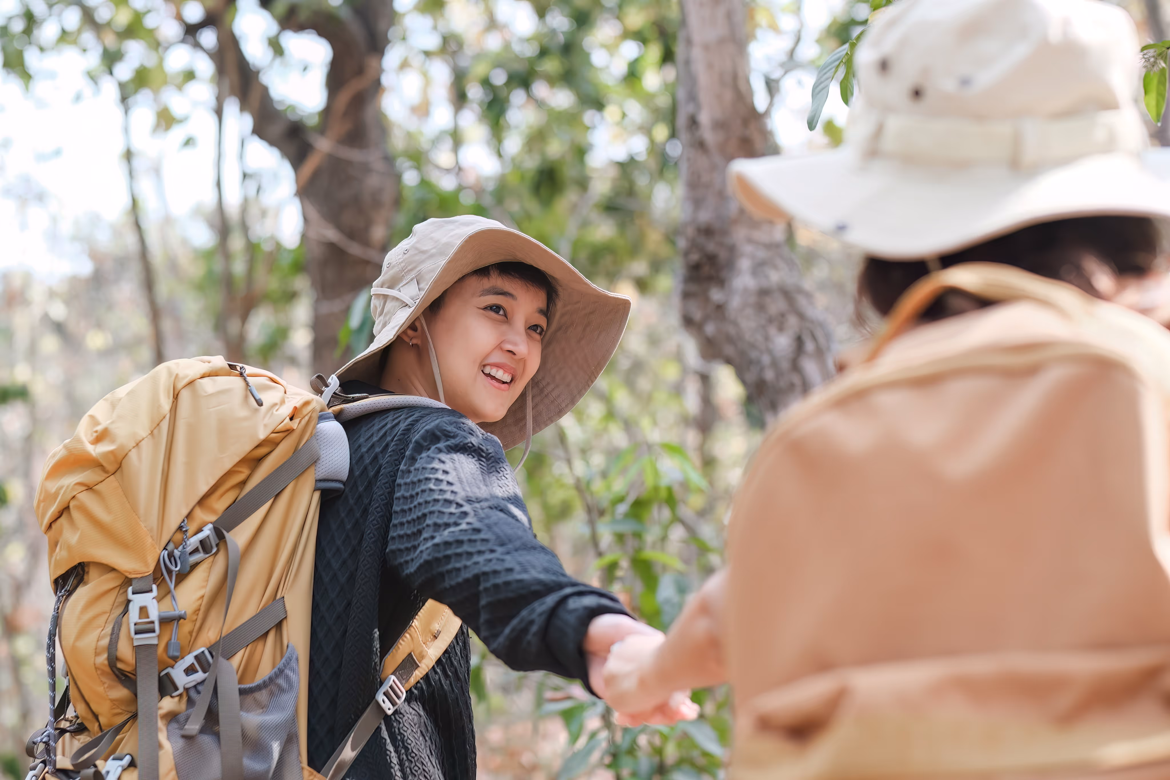 Smiling hiker with beige hat and backpack reaching out hand to another person in a forest.