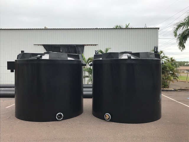 Two large black industrial storage tanks placed outdoors on pavement, with a white building and greenery in the background.
