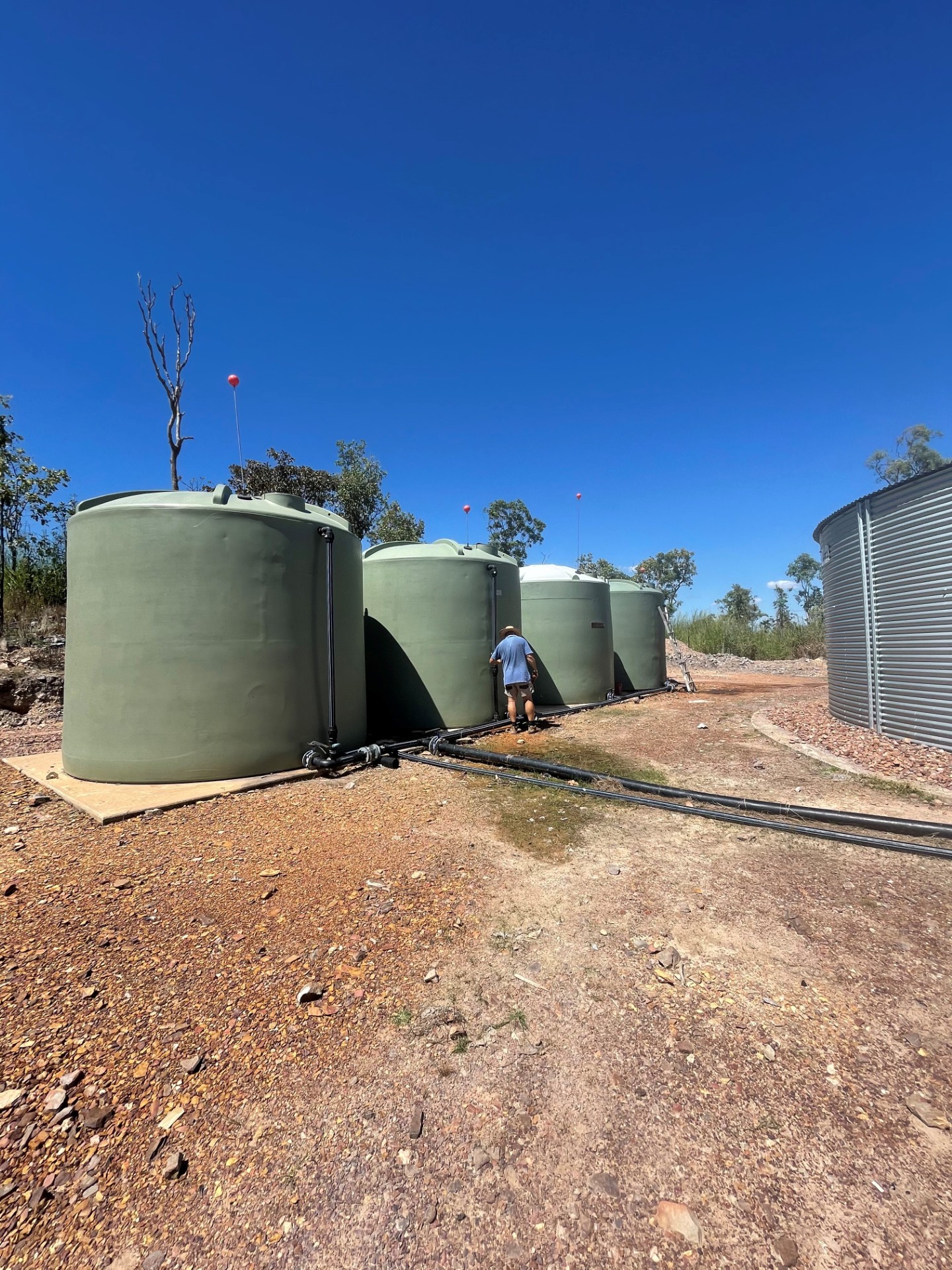 Four large green water storage tanks connected by black pipes on rocky ground under a clear blue sky with a person inspecting one tank.
