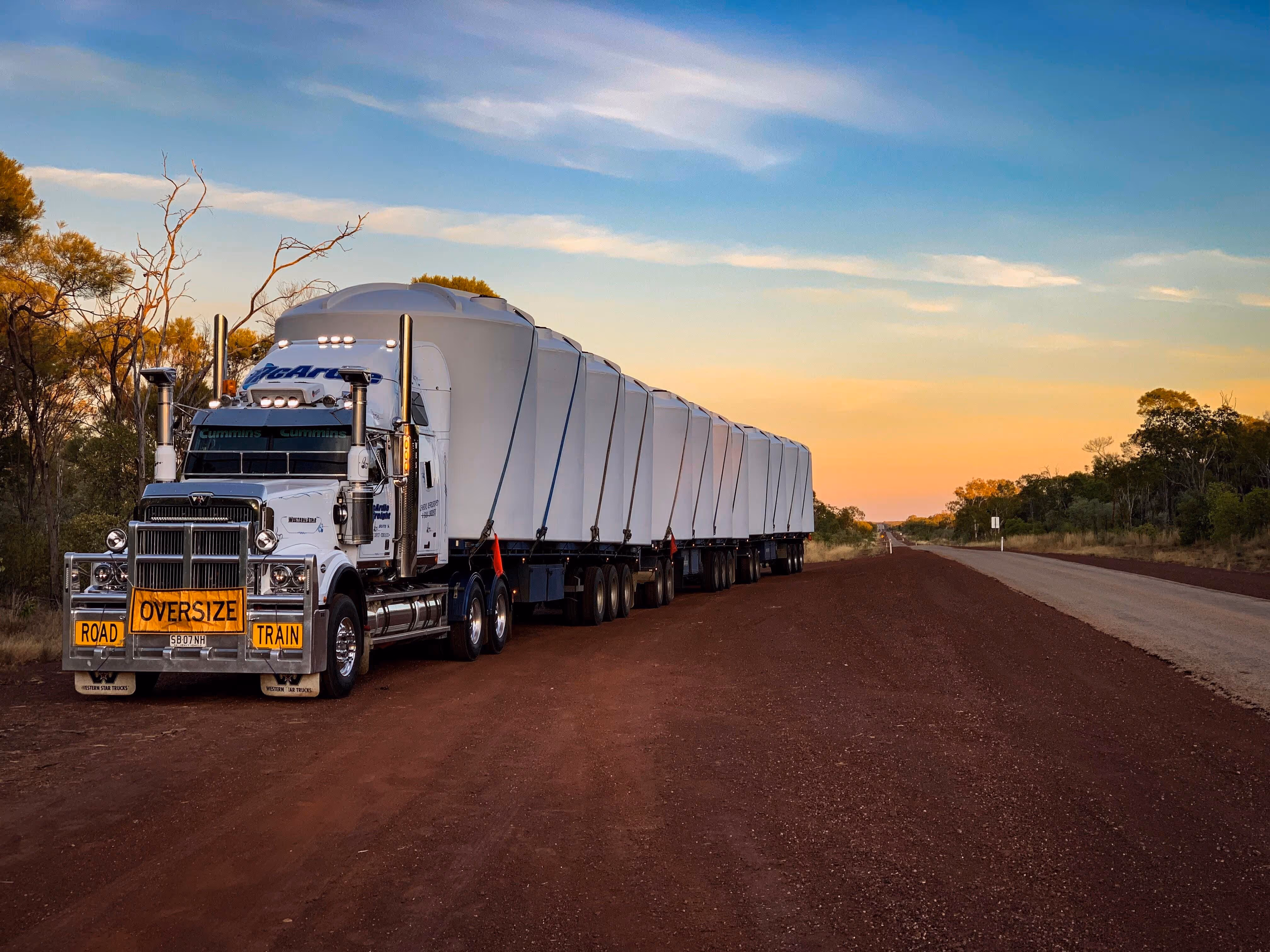 Large white road train truck with multiple trailers parked on a dirt road at sunset.