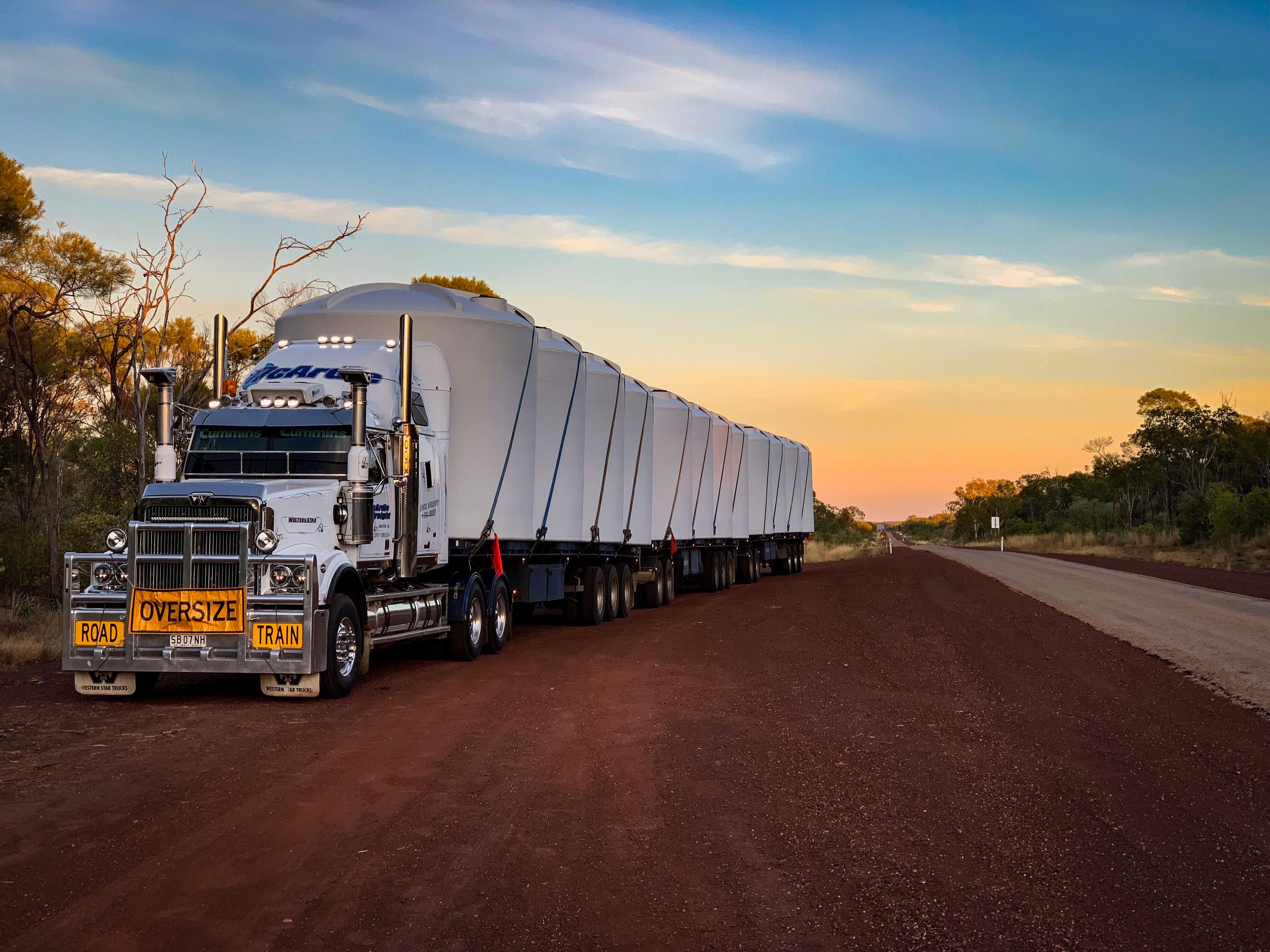 Large white road train truck with multiple trailers parked on a dirt road at sunset.
