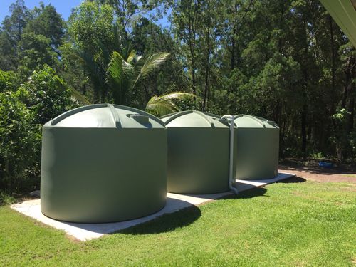 Three large green rainwater tanks on a concrete slab in a sunny yard with trees and grass.