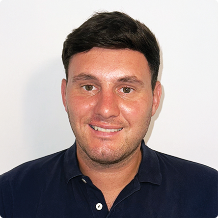 Man with short dark hair wearing a dark navy collared shirt, smiling against a plain white background.