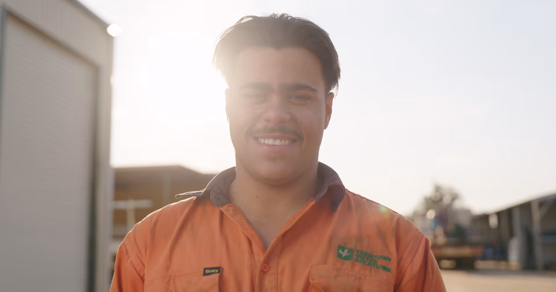 Smiling man in an orange work shirt standing outdoors with sunlight behind him.