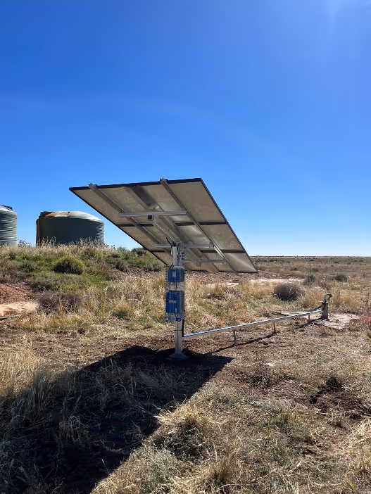 Rear view of a solar panel mounted on a single pole in a grassy field under a clear blue sky with two large water tanks in the background.