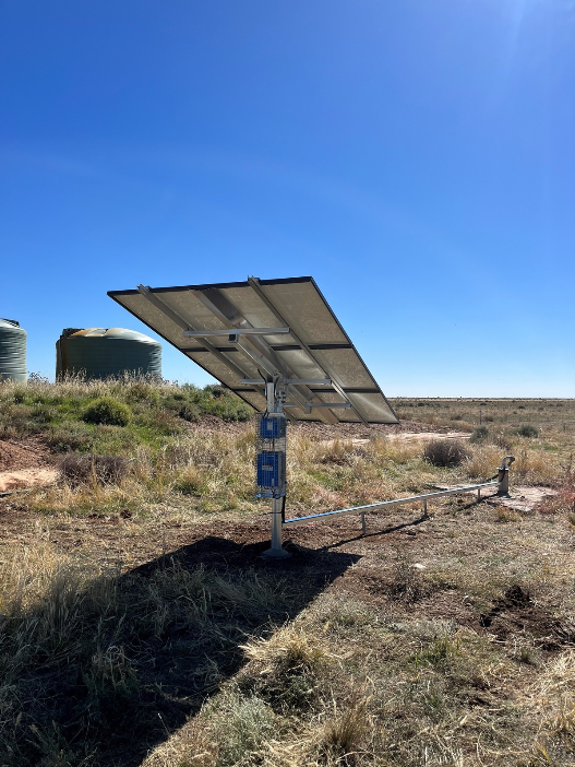Solar panel mounted on a pole in a grassy field with blue sky, connected to equipment on the ground and cylindrical water tanks in the background.