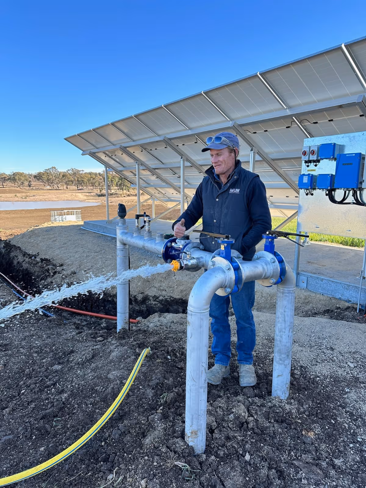 Man in outdoor workwear operating a water valve with flowing water and solar panels in the background under a clear blue sky.