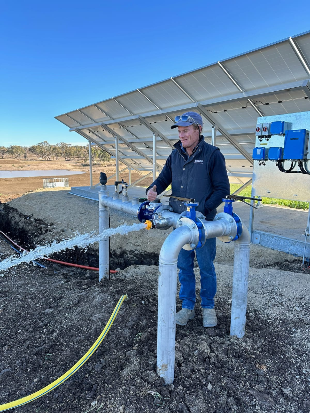 Man in outdoor workwear operating a water valve with flowing water and solar panels in the background under a clear blue sky.