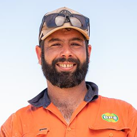 Smiling man with a beard wearing an orange shirt and a beige cap with sunglasses on top, against a clear sky.