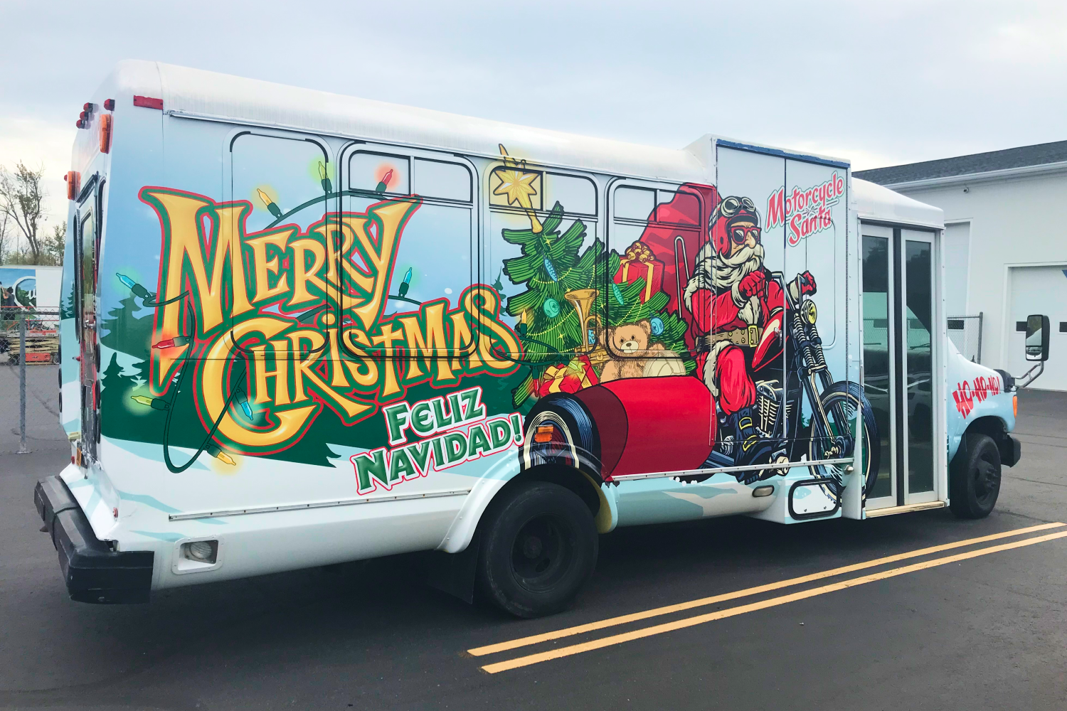 A medium sized bus with a wrap that features Santa riding a motorcycle delivering gifts and the words "Merry Christmas" in gold cursive lettering and wrapped in christmas lights.
