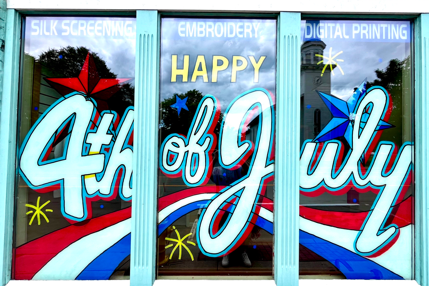 Window with stars, red, white, and bule stripes, and the words "Happy 4th of July" painted in white cursive letters.