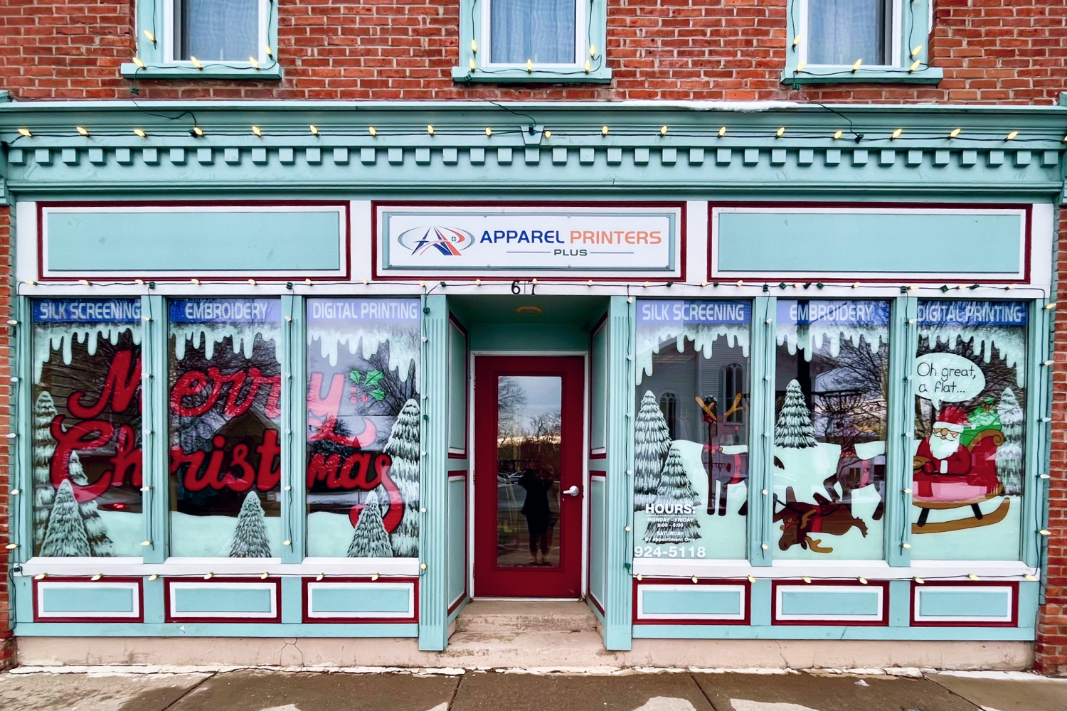 (Left Window) Window with white pine trees, snow, and the words "Merry Christmas" painted in red cursive letters. (Right Window) Three reindeer, one has fallen over, withe Santa in his sleigh saying "Oh great, a flat".