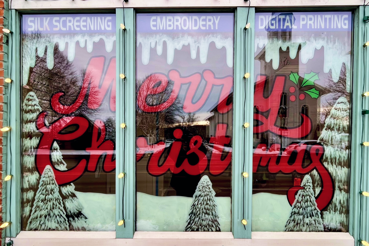 Window with white pine trees, snow, and the words "Merry Christmas" painted in red cursive letters.