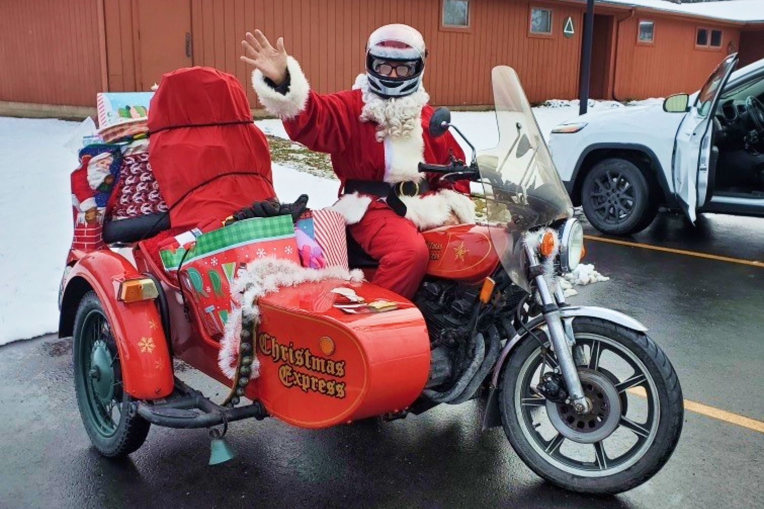 Man dressed in a Santa costume, waving while sitting on a christmas-themed motorcycle with a sidecar filled with gifts.