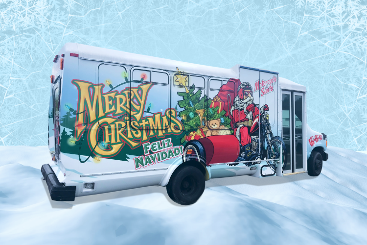 A medium sized bus with a wrap that features Santa riding a motorcycle delivering gifts and the words "Merry Christmas" in gold cursive lettering and wrapped in christmas lights.