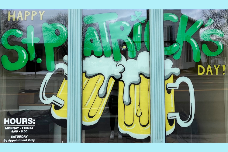 Window with two glasses of beer clinking and the words "Happy St. Patrick's Day" painted in green bubble letters. 