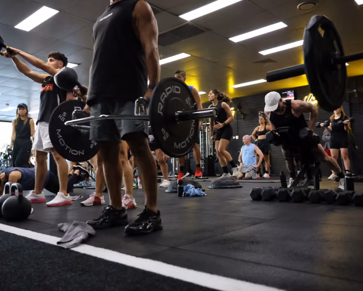 Several people lifting weights and exercising in a busy gym with black flooring and ceiling lights.
