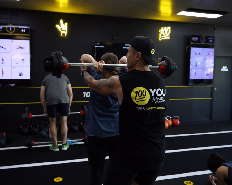 Man in black '100 YOU fitness' shirt coaching woman lifting barbell in a gym with screens and kettlebells.