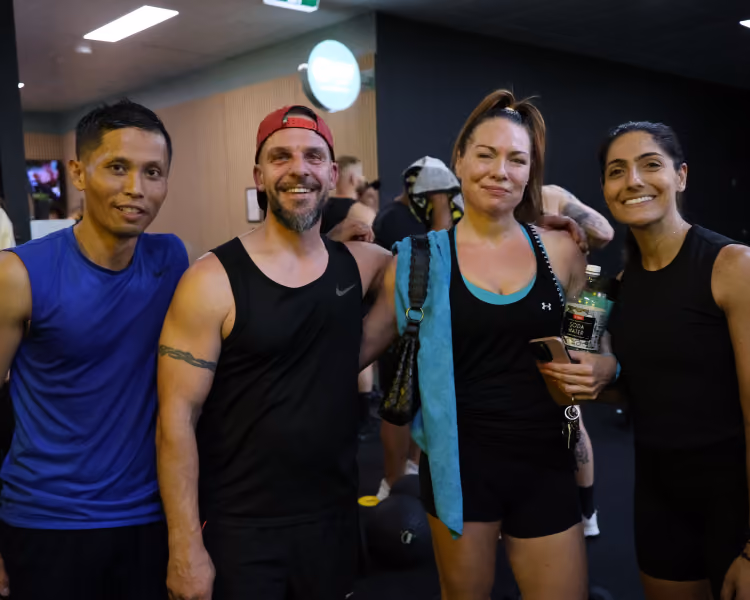 Four adults posing and smiling indoors, dressed in athletic wear, likely in a gym setting.
