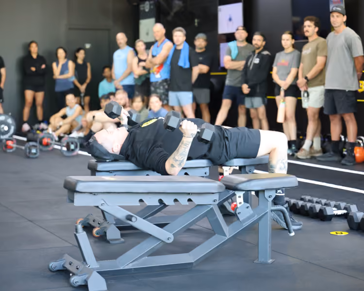 Man lifting dumbbells while lying on a bench with a crowd watching in a gym.