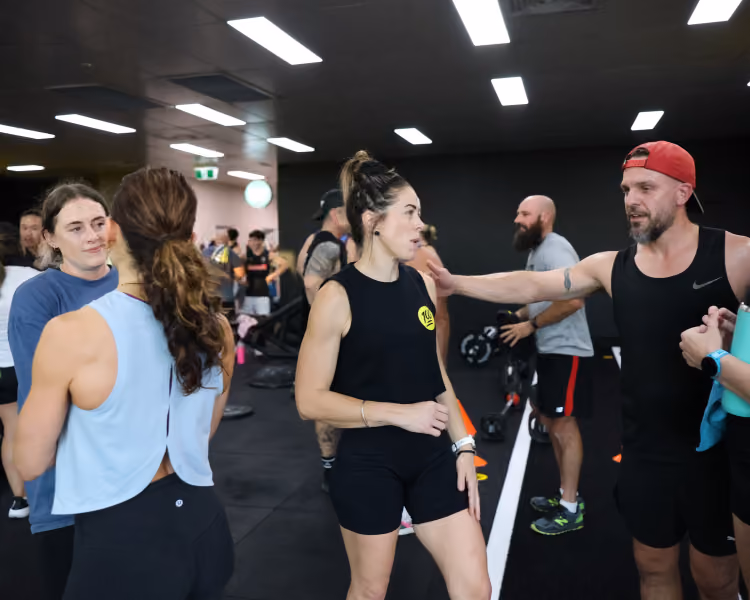 Group of adults in workout clothes talking and resting inside a gym with exercise equipment.