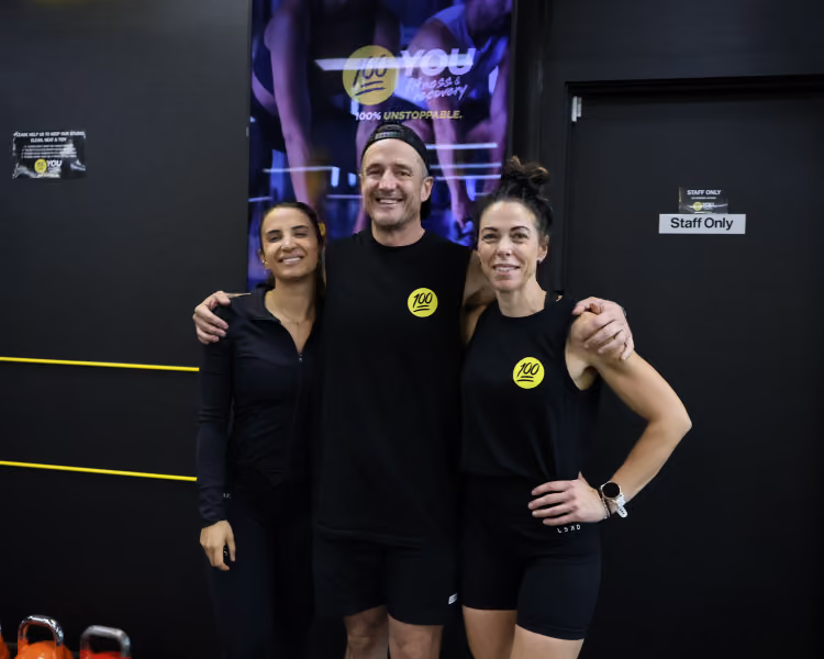 Three people smiling and posing together in a gym with black walls and workout equipment visible.