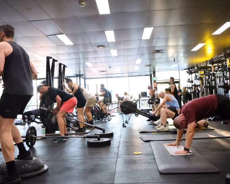 Multiple people exercising using weights, rowing machines, and mats in a spacious gym with large windows.